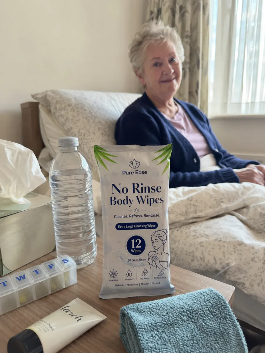 PureEase body wipes on a bedside table with care essentials and an older woman sitting up in bed.